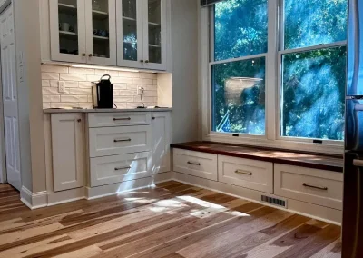 Bright kitchen with wood floors, white cabinets, a coffee maker, and a window bench under large windows.