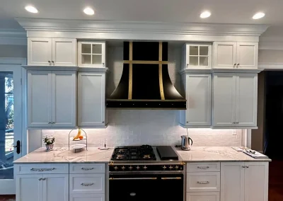 White kitchen with black and gold stove, matching vent hood, white cabinets, and marble countertops.