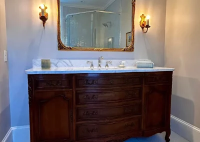 Ornate wood vanity with marble top, gold-framed mirror, and two wall sconces in a bathroom.