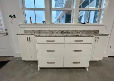 White cabinet with six drawers and granite countertop, placed in front of three windows in a room.