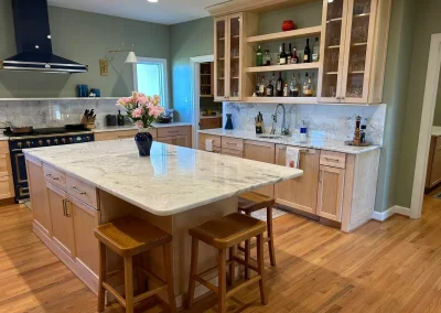 Sunny kitchen with wood cabinets, marble island, two stools, a black stove, and shelves with bottles.