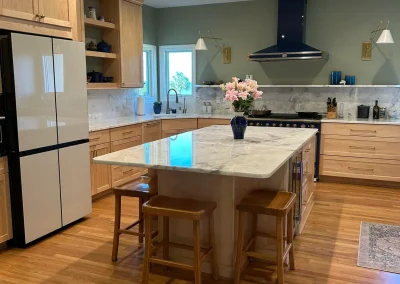 Modern kitchen with wood cabinets, marble island, stools, flowers in vase, and stainless steel appliances.