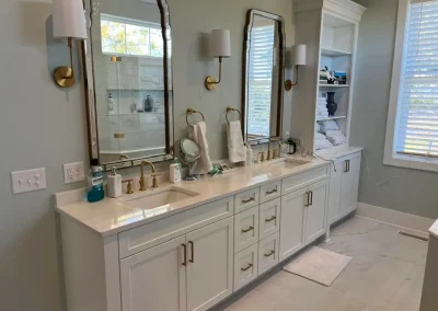 Double-sink bathroom vanity with mirrors, gold fixtures, shelves with towels, and natural light from windows.