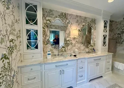 White double-sink bathroom vanity with ornate mirrors, floral wallpaper, and person reflected in mirror.