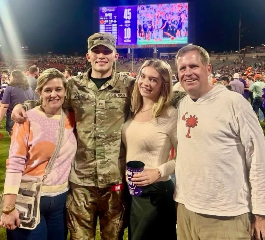 A smiling family poses with a U.S. Army soldier at a football stadium during a night game.