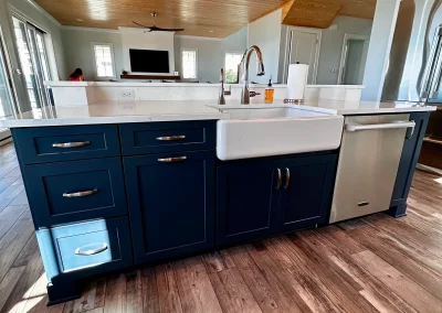 Modern kitchen island with navy cabinets, farmhouse sink, dishwasher, and wood flooring under natural light.