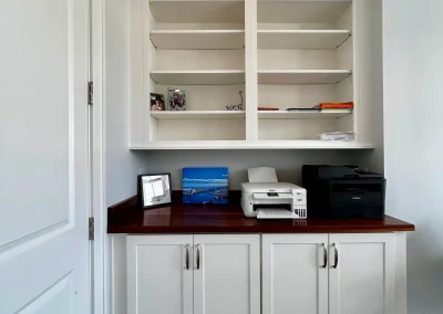 Built-in white office shelves and cabinets with a wooden countertop, printer, and framed photo on top.