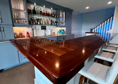 Home bar with a wooden countertop, shelves of bottles, and white bar stools beside a staircase.