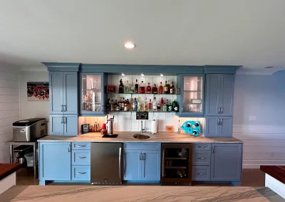 A home bar with blue cabinets, shelves of liquor bottles, glassware, and a small sink in the center.
