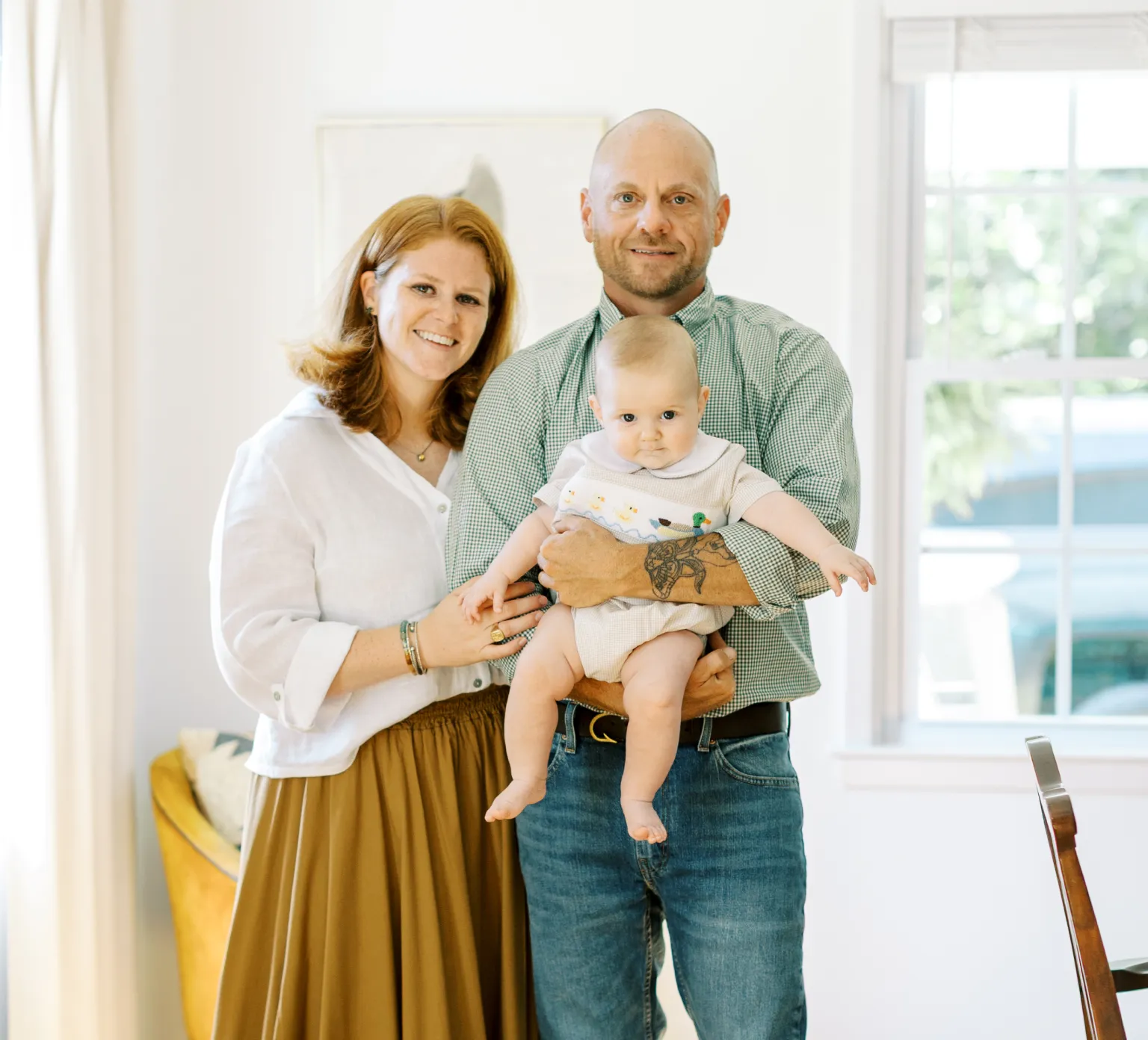 A smiling couple stands indoors, holding a baby, with sunlight streaming through a window behind them.