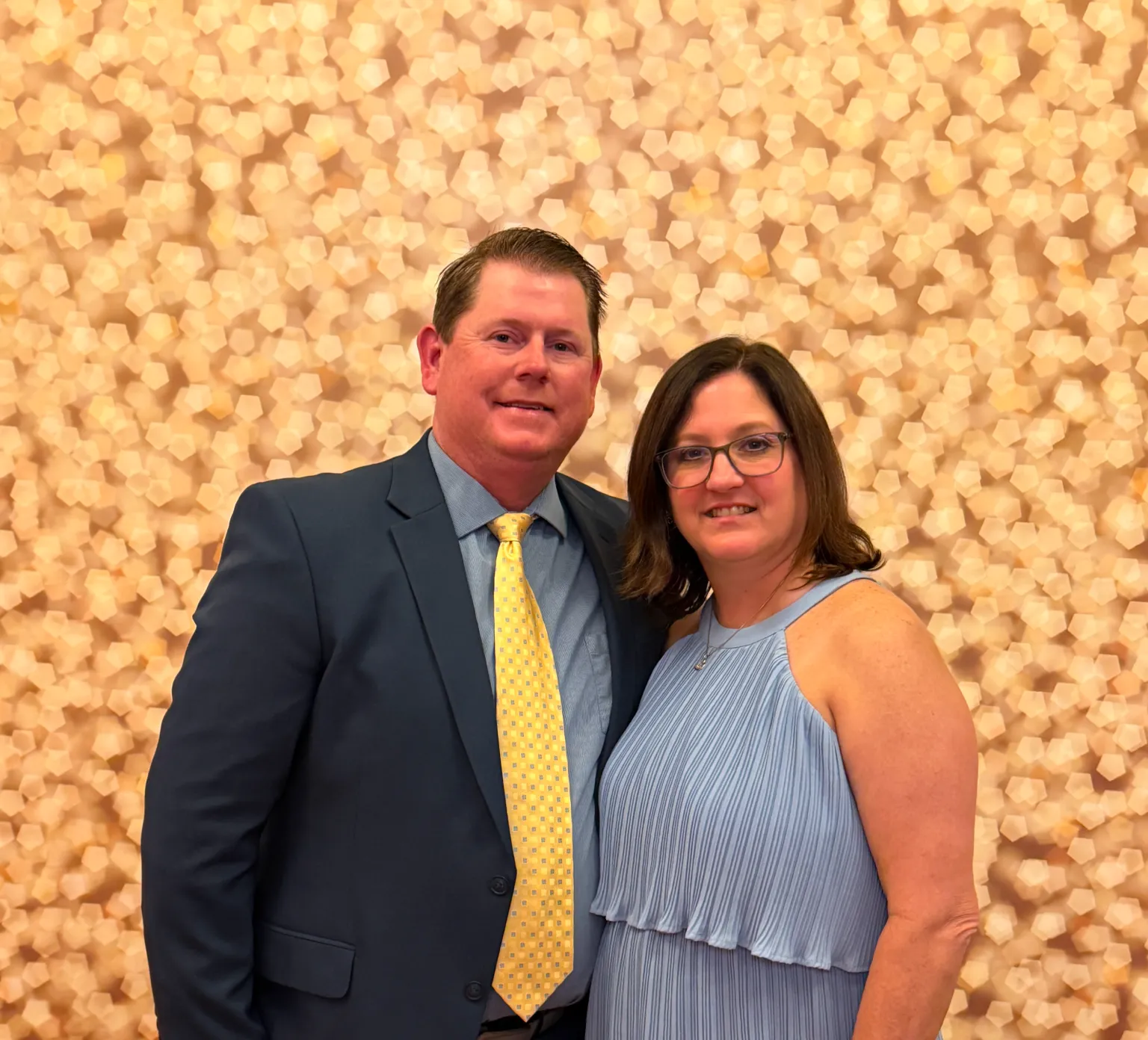 A man in a suit and a woman in a blue dress smile, standing in front of a light patterned backdrop.