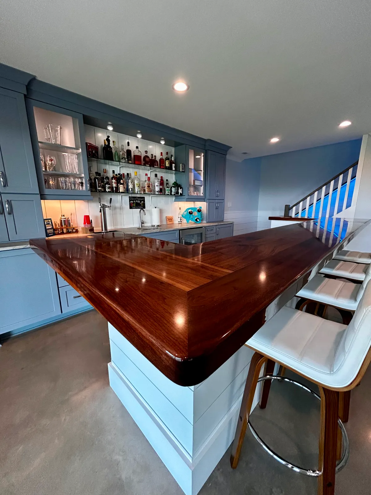 Modern home bar with dark wood counter, shelves with bottles, and white bar stools in a blue room.