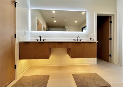 Modern bathroom with double sinks, wood cabinets, backlit mirror, and two mats on a light floor.