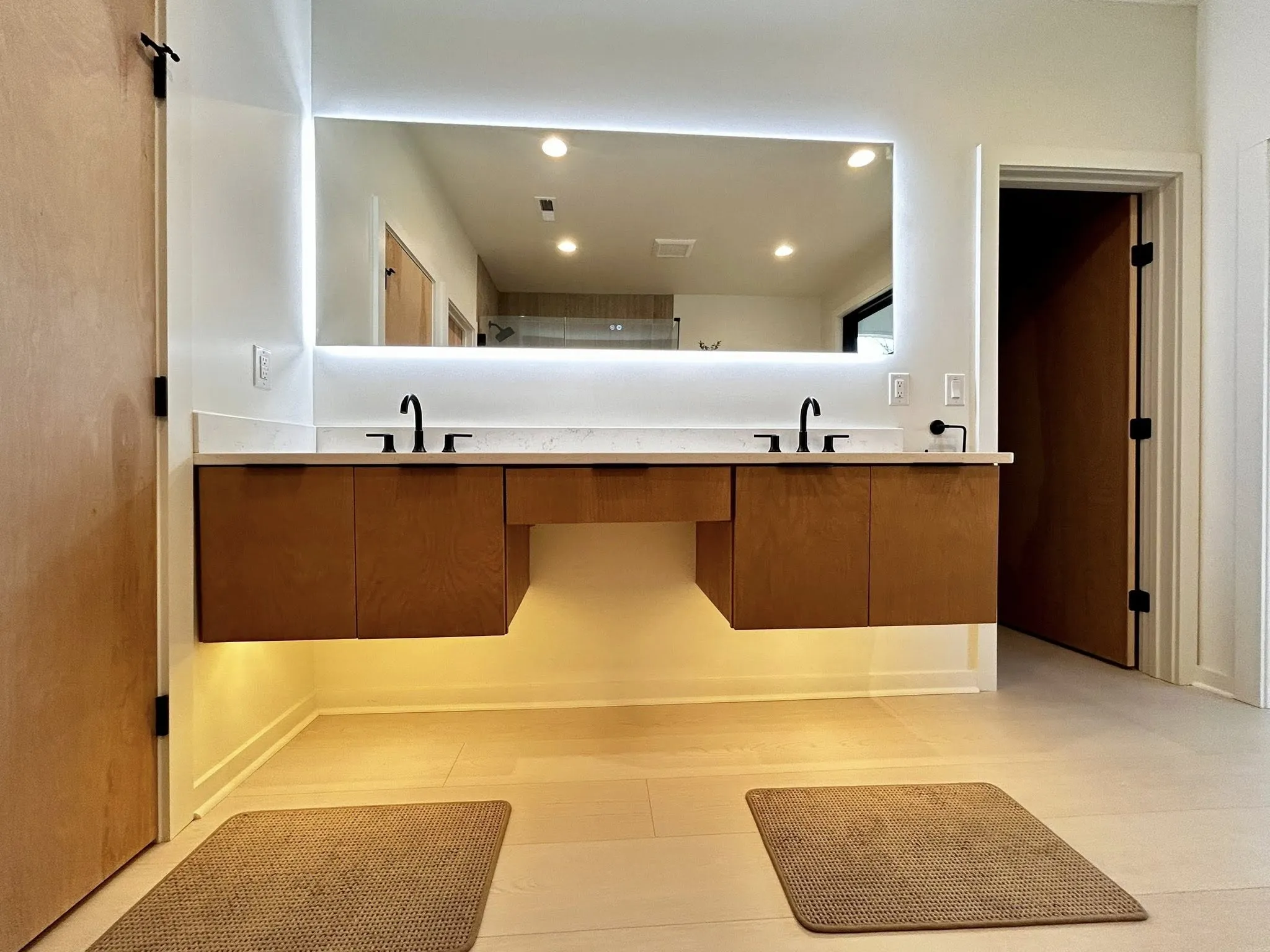 bathroom-c Modern bathroom with double sinks, wood cabinets, backlit mirror, and two mats on a light floor.