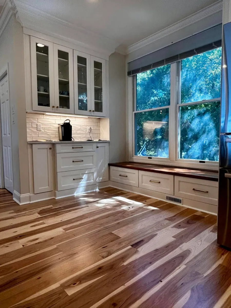 Bright kitchen corner with wood floors, white cabinets, and a window bench seat overlooking trees.