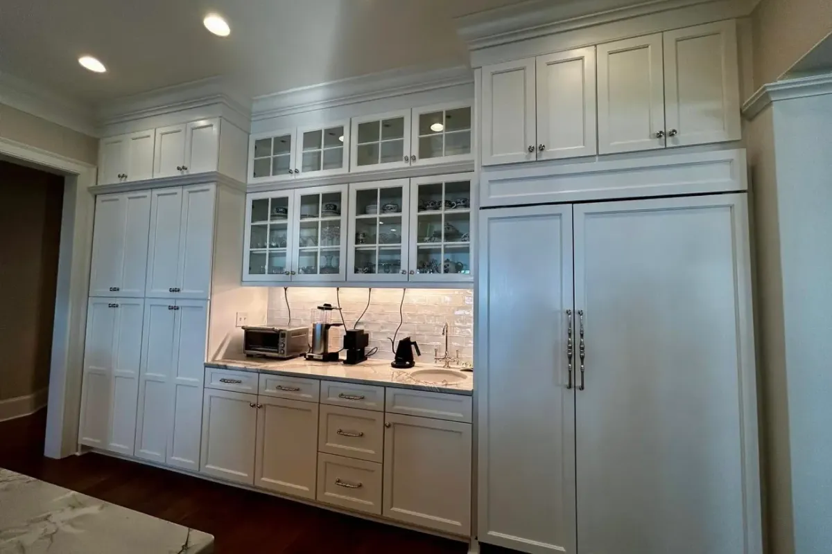 White kitchen cabinets with glass doors, a sink, and countertop appliances under bright ceiling lights.