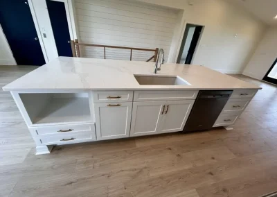 White kitchen island with sink, dishwasher, cabinets, drawers, and open shelves on light wood floor.