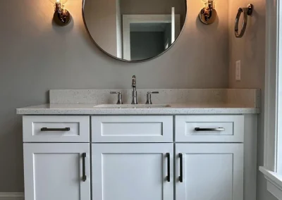 White bathroom vanity with a round mirror, two wall lights, and a window to the right.