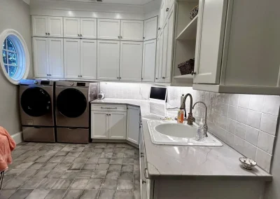 Modern laundry room with white cabinets, sink, washer, dryer, tiled floor, and round window.