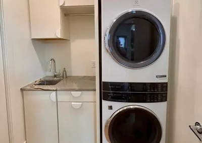 Stacked washer and dryer next to a small sink and cabinet in a laundry nook with white walls.