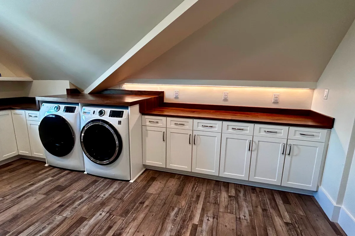 Modern laundry room with washer, dryer, white cabinets, wood countertops, and sloped ceiling.