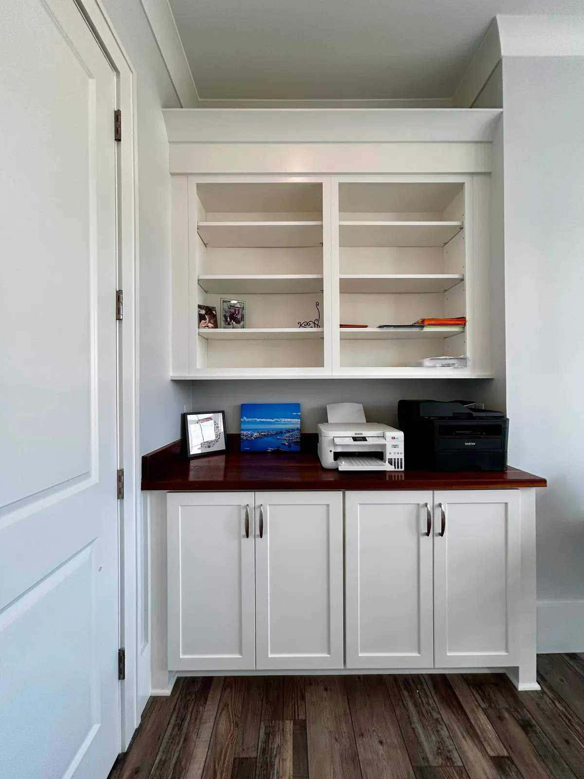 White built-in shelves above a wooden countertop with printers, photos, and cabinets below on a wood floor.