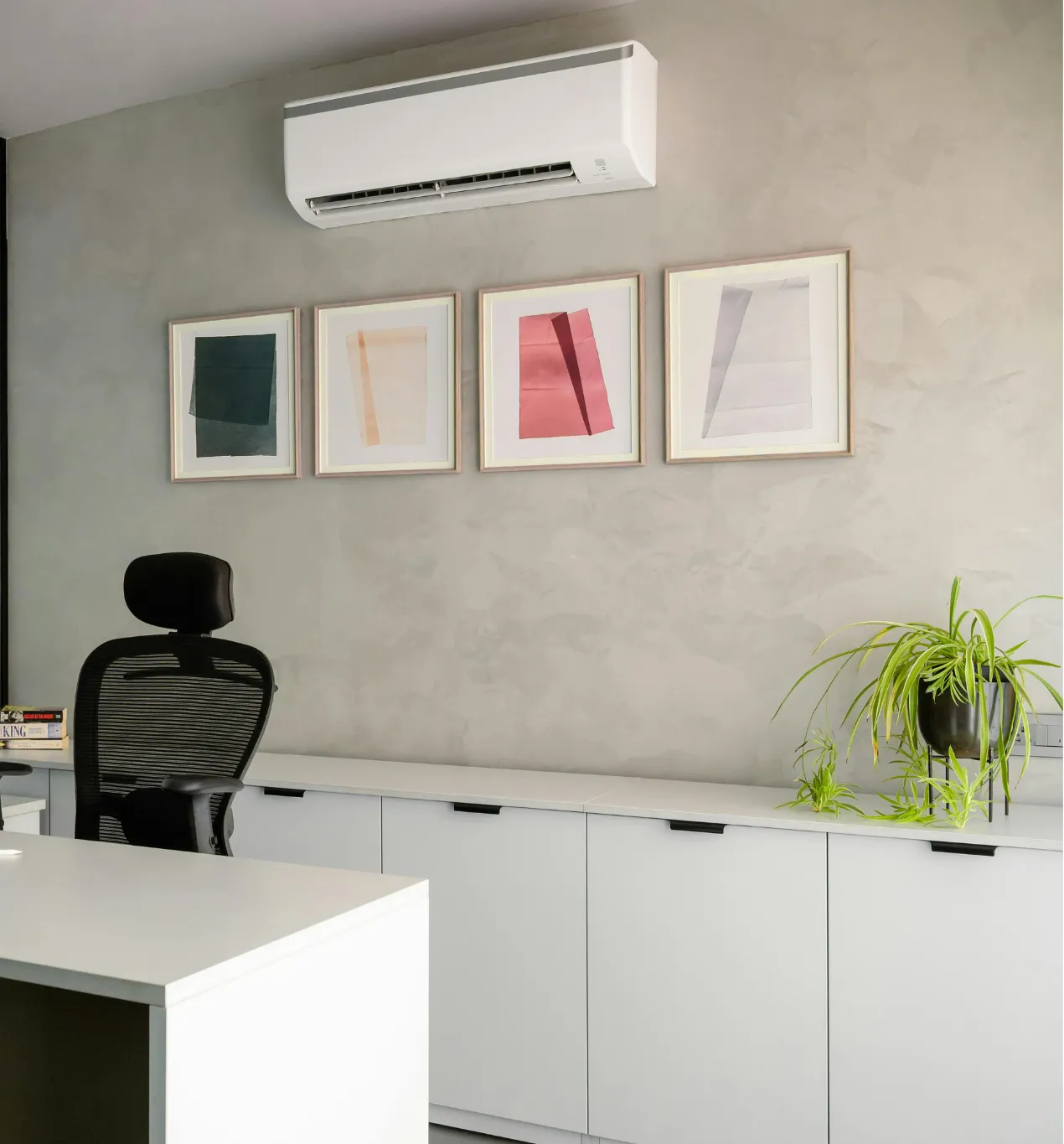 Modern office with a desk, black chair, plant, air conditioner, and four abstract framed artworks on the wall.
