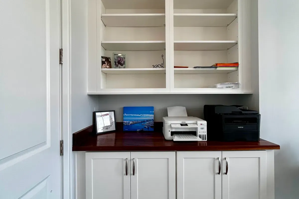Office nook with white shelves, framed photos, a printer, and a copier on a wooden countertop.