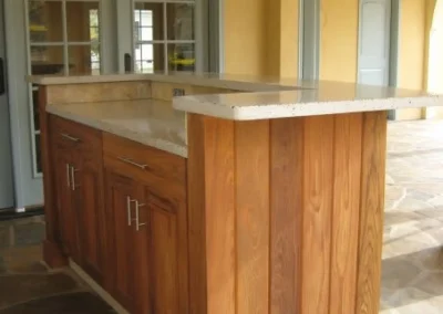 Wooden outdoor kitchen island with a stone countertop on a tiled patio, in front of double doors.