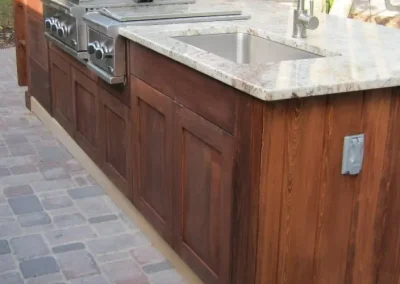 Outdoor kitchen with a built-in grill, stainless steel sink, and wooden cabinetry on a stone patio.