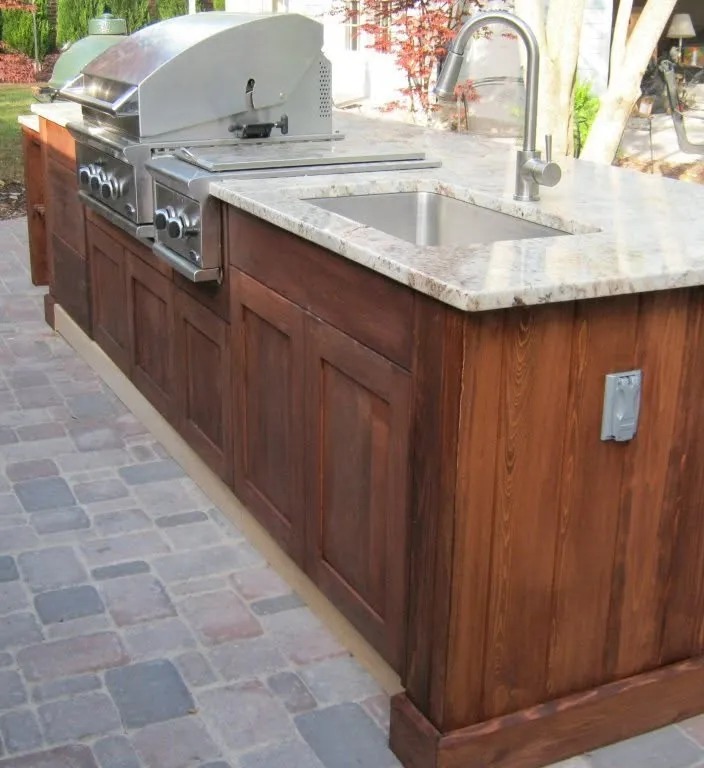 Outdoor kitchen with a built-in grill, stainless steel sink, and wooden cabinetry on a stone patio.