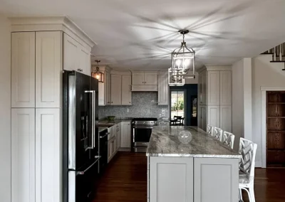 Modern kitchen with white cabinets, black fridge, island with marble top, and three white chairs on wood floor.