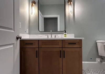 Modern bathroom with wood vanity, square mirror, wall lights, patterned tile floor, and a toilet on the right.