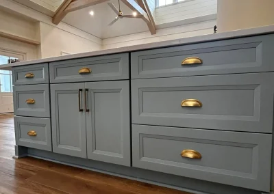 Gray kitchen island with gold handles and drawers, set on a wooden floor in a bright room with high ceilings.