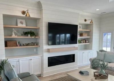 A modern living room with a wall-mounted TV, fireplace, shelves, and cabinets with decor and plants.