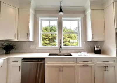 Bright modern kitchen with white cabinets, sink under double window, and greenery visible outside.