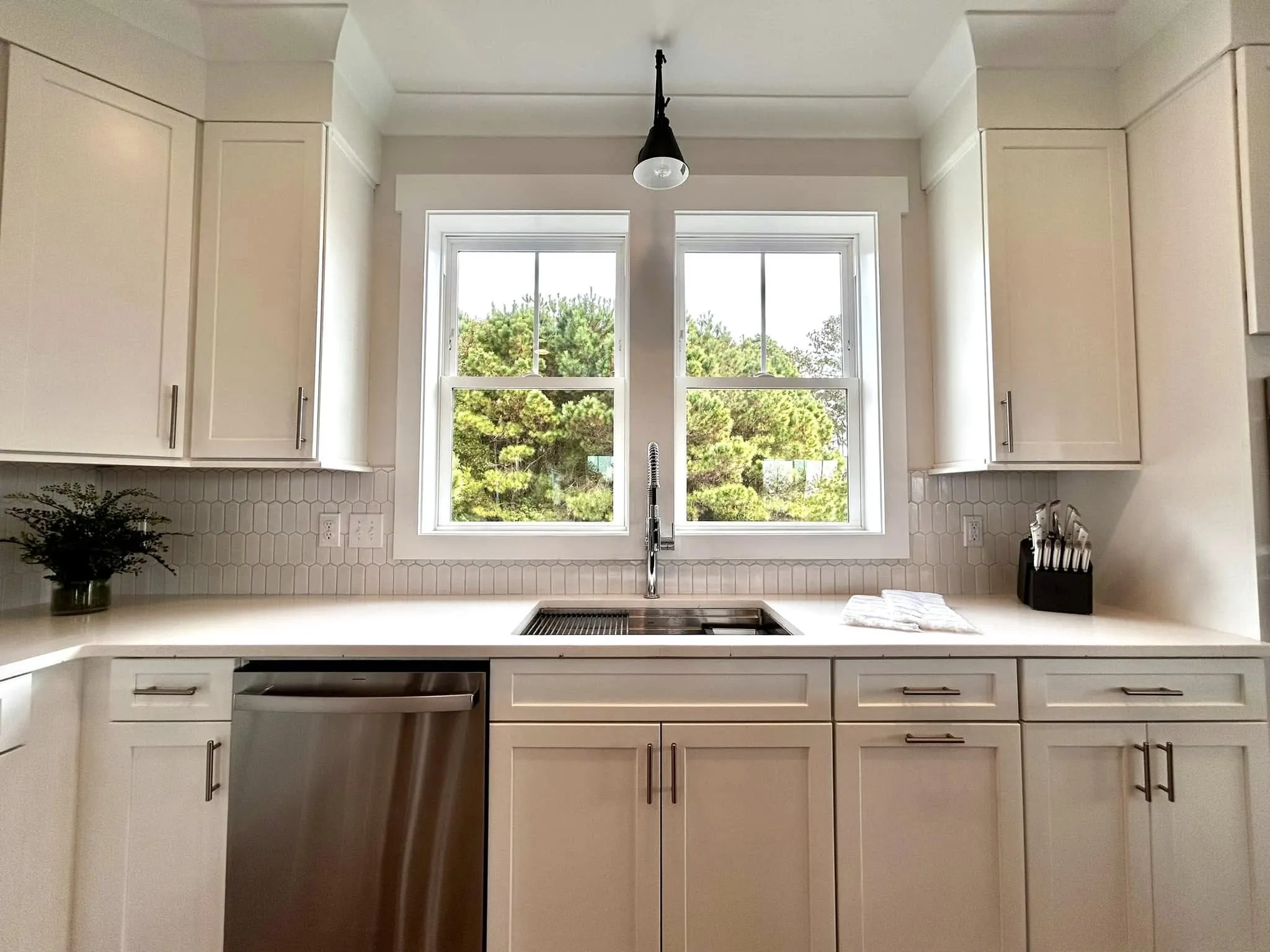Bright modern kitchen with white cabinets, sink under double window, and greenery visible outside.