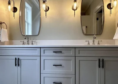 Modern bathroom with double sinks, oval mirrors, light fixtures, gray cabinets, and towel rings on each side.