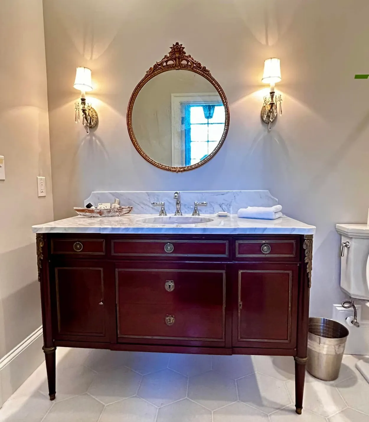 vanity-2 Elegant bathroom vanity with round mirror, marble countertop, and wall sconces; toilet and trash bin nearby.